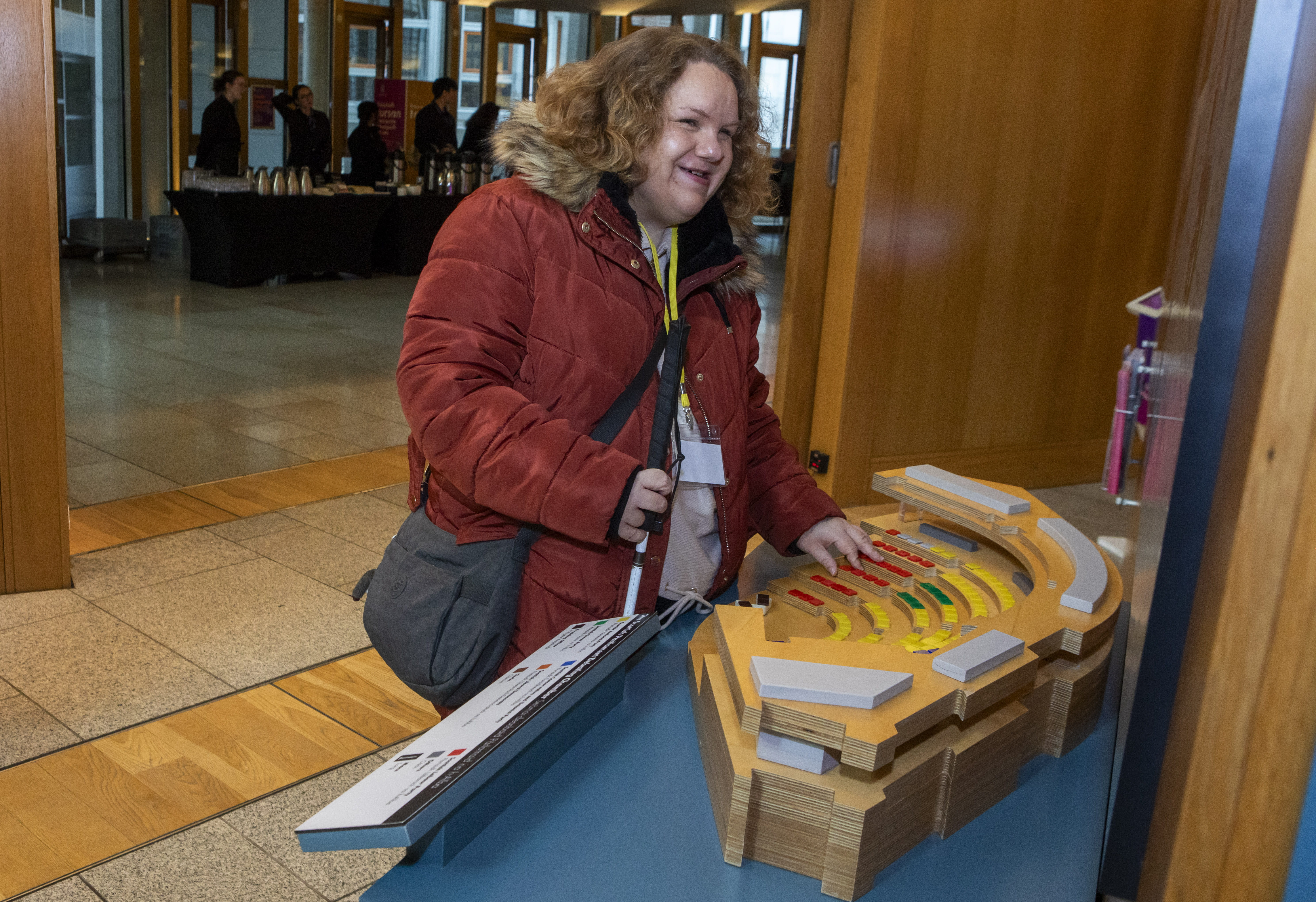 Individual with white cane explores a tactile model of a parliamentary chamber, featuring raised and color-coded seating in a public building.