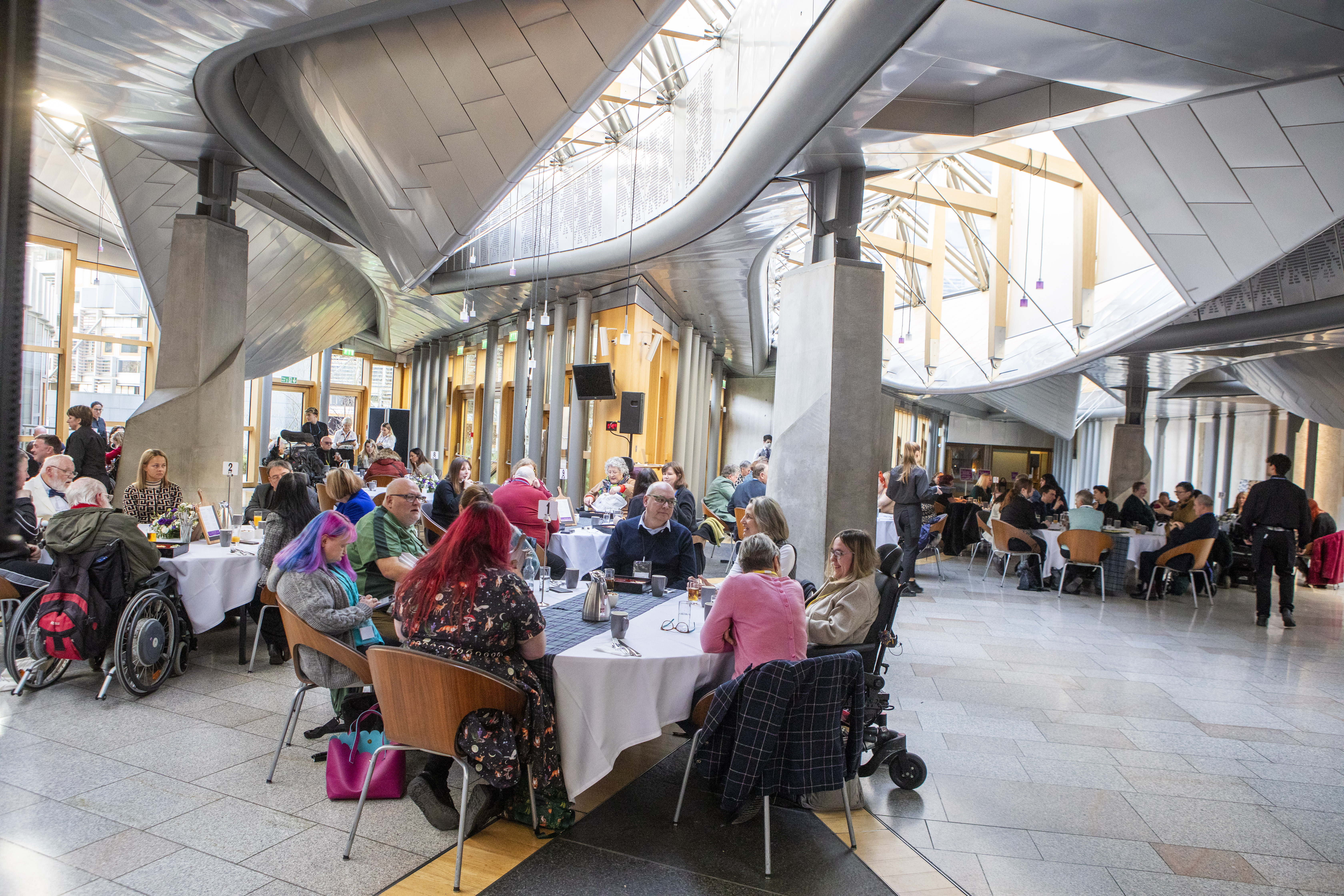 Large contemporary hall with high metal-and-glass ceiling, filled with round tables and diverse attendees, including wheelchair users, engaged in dining and conversation