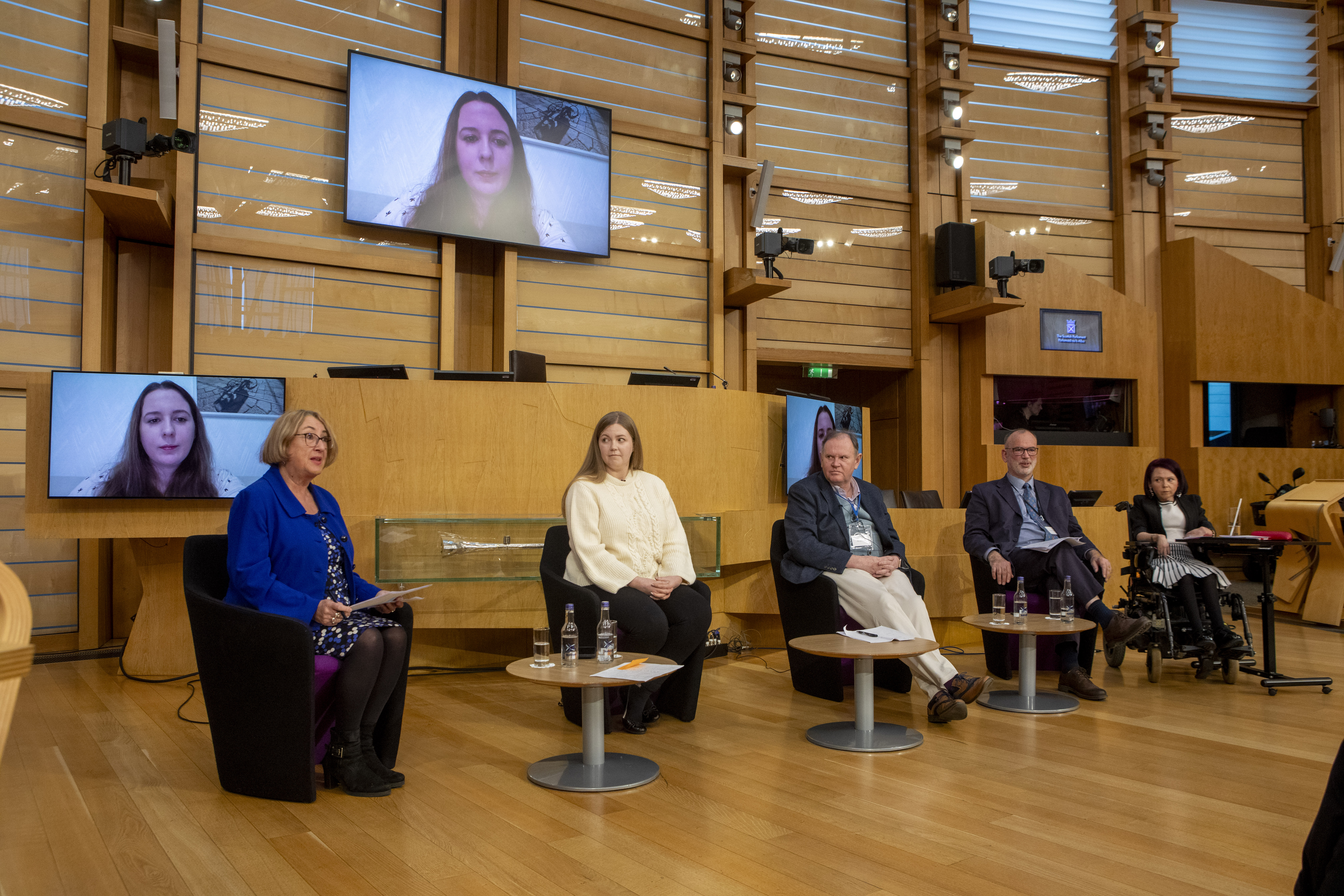 Five panelists seated on stage with water bottles and papers; hybrid event includes remote speaker on screen and visible accessibility accommodations