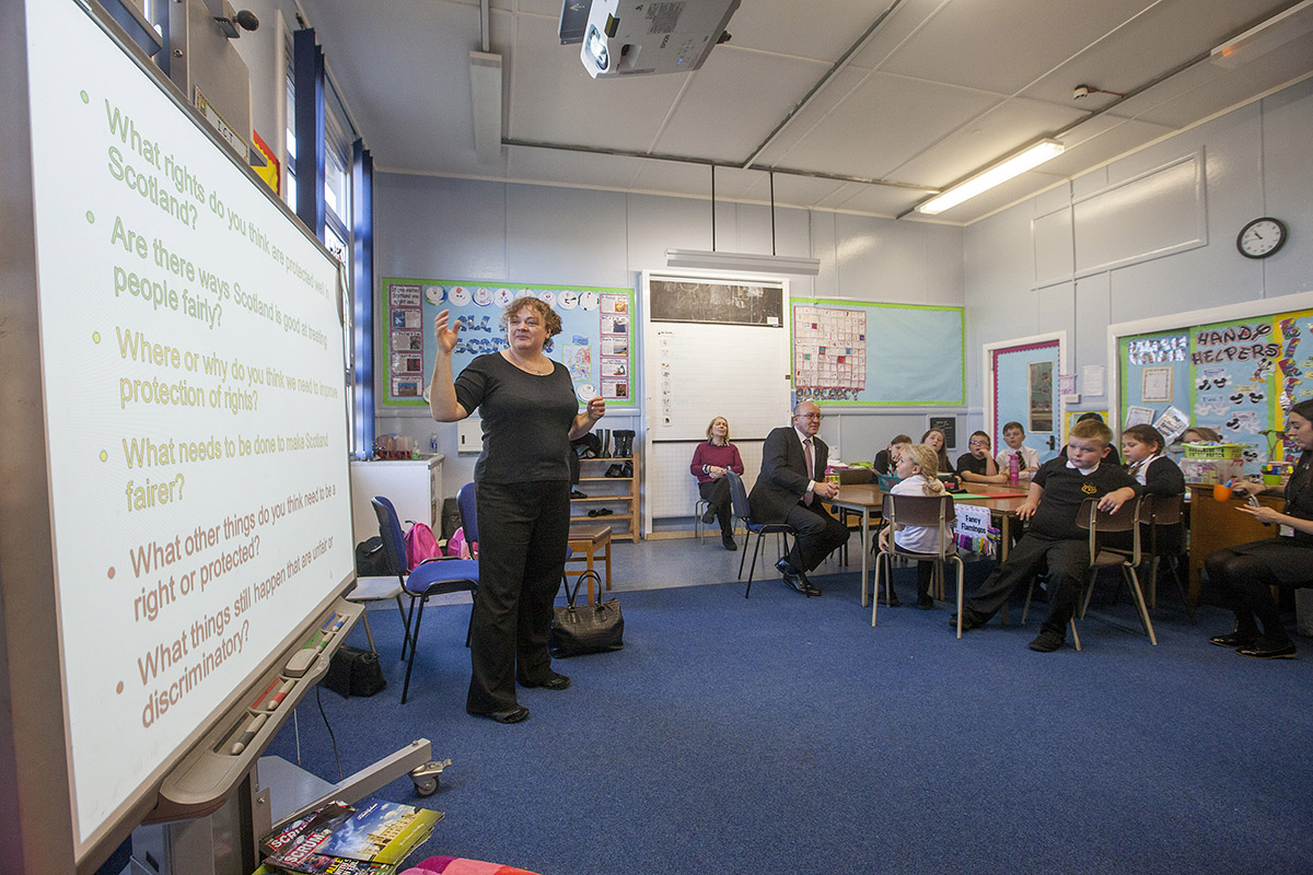 A teacher stands in front of a projector screen in a classroom of pupils