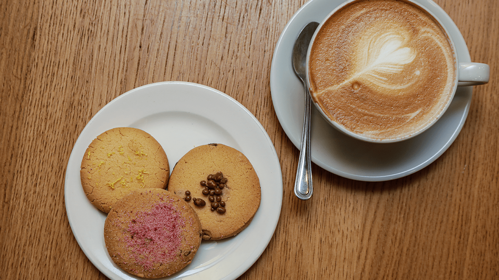 Cappuccino with heart-shaped latte art next to a white plate holding three assorted shortbread biscuits on a wooden table