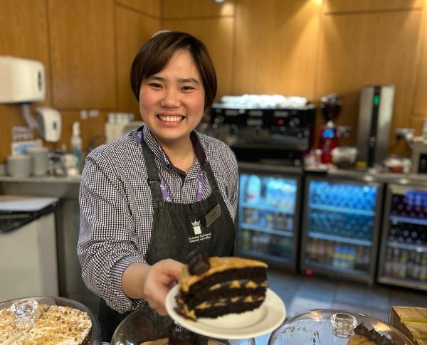 A smiling staff member at the cafe holds out a piece of chocolate cake on a plate