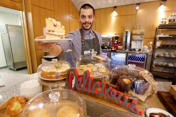 a café staff member holds up a slice of cake above a counter covered in many other cakes and pastries